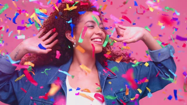 A happy smiling young woman wearing a denim jacket under a cascade of colorful confetti falling in slow motion.