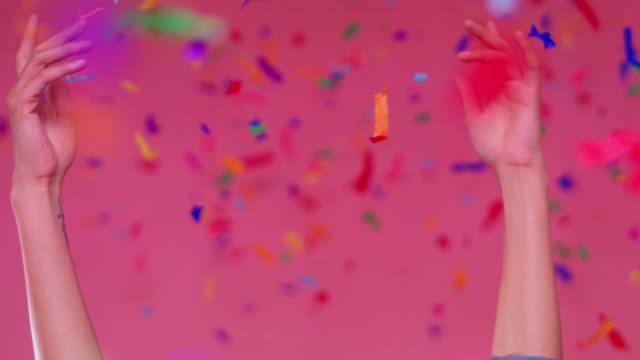 Close up shot of a young smiling woman with her arms extended to the top while confetti falls over a pink background.