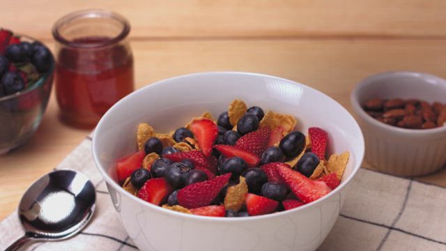 A continuous stream of pristine white milk falling and splashing into a bowl full of corn flakes, blueberries and strawberries.