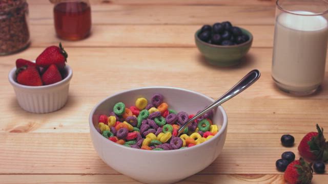 A continuous stream of milk falling into a bowl brimming with colorful cereal rings on a beautiful rustic table decorations with a plate of fresh strawberries, honey and a glass of milk.