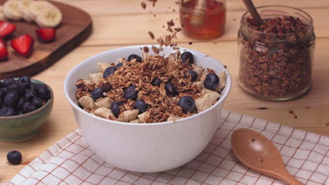 Granola grains fall down in slow motion into a bowl brimming with cereal and blueberries.