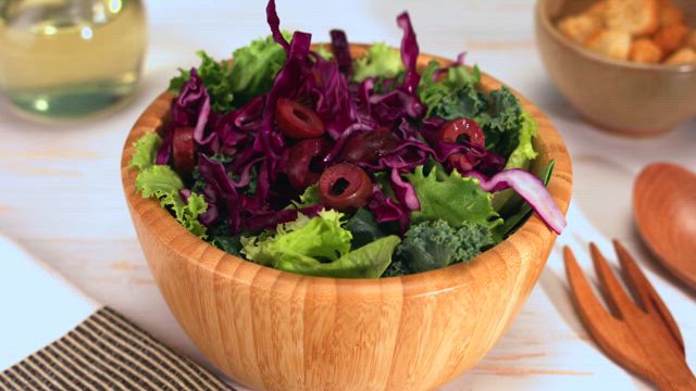 Red cabbage slices falling into a rustic wooden bowl brimming with green lettuce over a table with a white tablecloth.