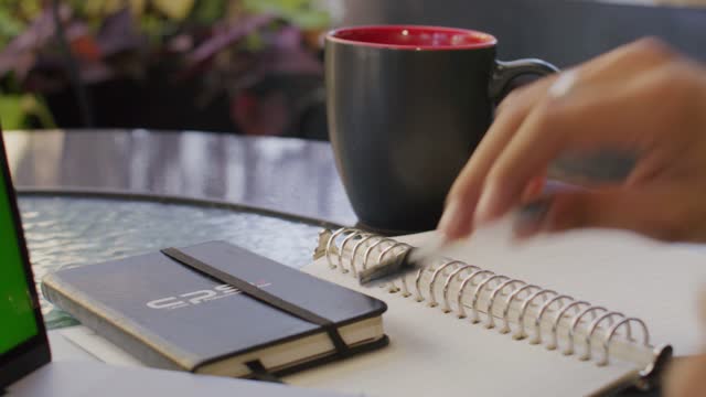 The hands of a man taking notes with a pen on a cafe table with a black coffee cup in the back.