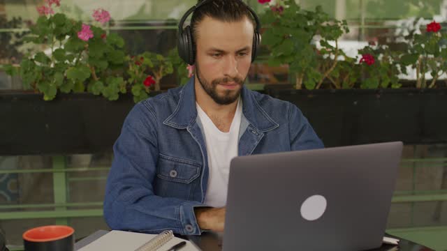 A young entrepreneur working on his laptop on a cozy cafe table vibing to the music from his headphones.