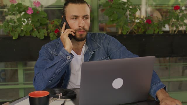 A young man talking on his mobile phone in front of a laptop computer.