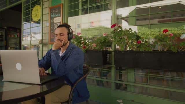 A young man wearing headphones sits in front of his laptop computer at the cafe table then takes a sip of coffee.