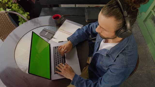 A man sitting on a cafe table types on his laptop computer showing a green screen then takes a pause to take a sip of delicious coffee.