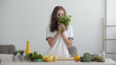 Young Smiling Woman Sniffing Fresh Greens Standing at the Table in Modern Kitchen.