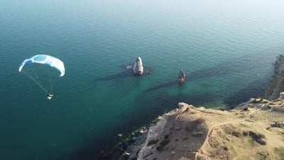 Aerial Drone View of a Man Flying a White and Blue Paraglider Over a Hill and Trees to the Sea Waves.