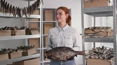 Small Business Portrait of Young Smiling Woman with Big Salted Fish in Her Hands Stands in Warehouse.