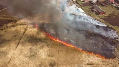 Aerial view of a field with dry grass set on fire with orange flames and high column of smoke..